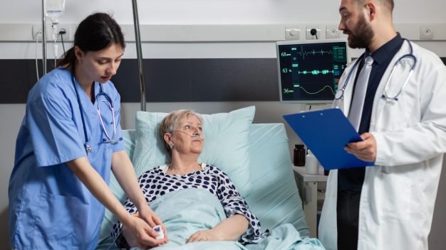 Medical nurse attaching oxymeter on senior woman patient, laying on hospital bed discussing with doctor about diagnosis and treatment while she breaths with help from oxygem mask.