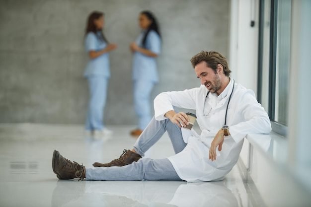 stressed-mature-doctor-sitting-floor-near-window-while-having-quick-coffee-break-hospital-hallway-covid-19-pandemic_360066-5554